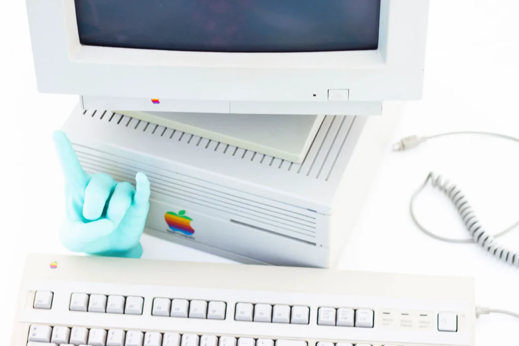 Computer Apple vintage con monitor CRT e tastiera, accanto una mano scultorea azzurra che fa il segno del rock